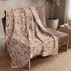 May include: A throw blanket featuring a repeating pattern of watercolor-style leaves and branches in shades of rust, orange, brown, and muted green on a cream-colored background, draped over a brown upholstered chair.