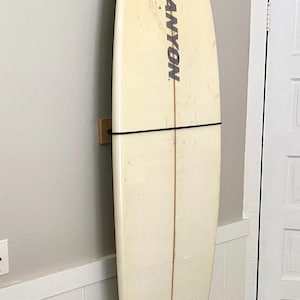 May include: Off-white surfboard with the word "CANYON" in black lettering. The surfboard has three black fins and is mounted on a wooden rack against a light gray wall. A black strap secures the board.