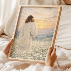 May include: A framed print depicts a figure in a white robe holding another person, set against a field of white flowers and a sunset. The artwork is in a light wooden frame, held by a person.