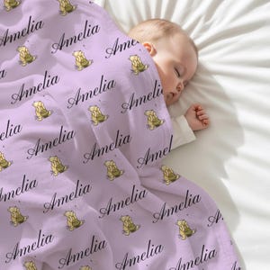May include: A light purple baby blanket with the name "Amelia" in a cursive font. The blanket has a repeating pattern of a cartoon bear holding a cake. A baby is sleeping under the blanket.