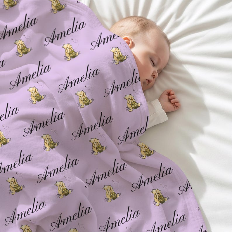 May include: A light purple baby blanket with the name "Amelia" in a cursive font. The blanket has a repeating pattern of a cartoon bear holding a cake. A baby is sleeping under the blanket.