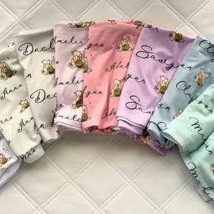 May include: A collection of personalized baby pajamas in various pastel colors, including lavender, pink, and mint green. Each set features a cartoon bear illustration and a name in cursive script. The pajamas are folded neatly.