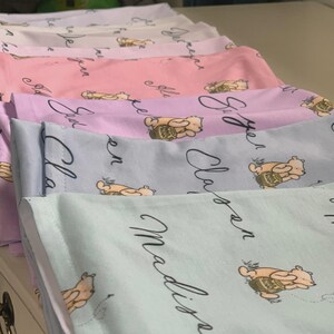 May include: A stack of pastel-colored baby blankets. Each blanket features a repeating pattern of a cartoon bear with a honey pot and a name in cursive script. The colors include light pink, lavender, blue, and green.