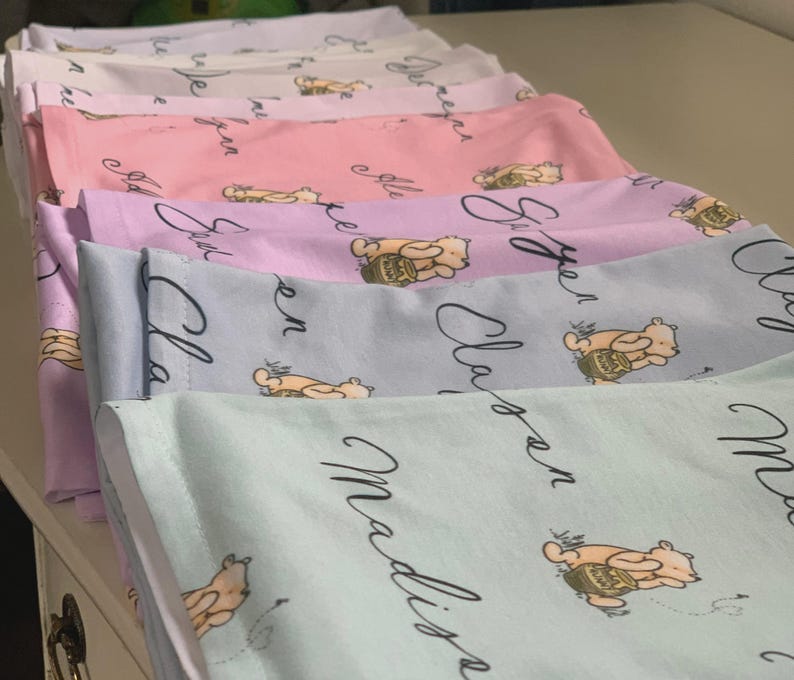 May include: A stack of pastel-colored baby blankets. Each blanket features a repeating pattern of a cartoon bear with a honey pot and a name in cursive script. The colors include light pink, lavender, blue, and green.