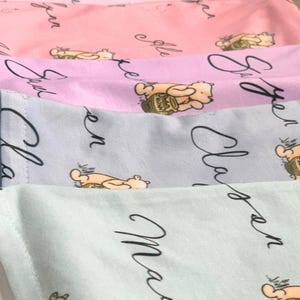 May include: A stack of pastel-colored fabric items, each featuring a repeating pattern of a cartoon bear with a honey pot. The fabric has cursive text in black. Colors include pink, lavender, and light blue.