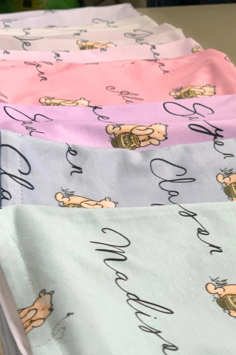 May include: A stack of pastel-colored fabric items, each featuring a repeating pattern of a cartoon bear with a honey pot. The fabric has cursive text in black. Colors include pink, lavender, and light blue.