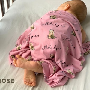 May include: A pink baby blanket with a repeating pattern of a cartoon bear holding a honey pot and the name "Adelyn" in cursive. The blanket is draped over a baby lying on a white surface. The word "ROSE" is in the bottom left corner.