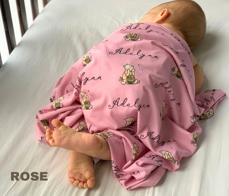 May include: A pink baby blanket with a repeating pattern of a cartoon bear holding a honey pot and the name "Adelyn" in cursive. The blanket is draped over a baby lying on a white surface. The word "ROSE" is in the bottom left corner.