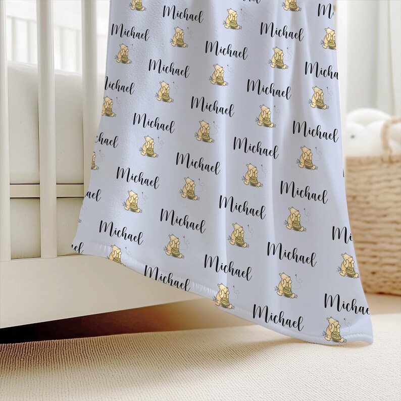 May include: A light blue baby blanket with a repeating pattern of the name "Michael" in black script and a cartoon bear with a honey pot. The blanket is draped over a white crib.
