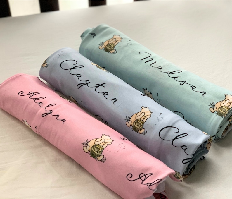 May include: Three personalized baby blankets with Winnie the Pooh characters. The blankets are rolled up and stacked on top of each other. The top blanket is light blue with the name "Madison" written on it. The middle blanket is light purple with the name "Clayton" written on it. The bottom blanket is pink with the name "Adelynn" written on it.