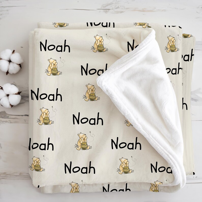 May include: A cream-colored baby blanket featuring the name "Noah" in black, repeated across the surface. The blanket also has a cartoon illustration of a bear holding a honey pot. The blanket is folded, revealing a soft, white interior.