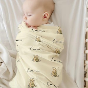 May include: A sleeping baby wrapped in a light yellow blanket. The blanket is patterned with a cartoon bear, honey pot, and the name "Emma" in black script. The baby is in a white crib.