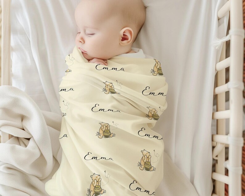 May include: A sleeping baby wrapped in a light yellow blanket. The blanket is patterned with a cartoon bear, honey pot, and the name "Emma" in black script. The baby is in a white crib.
