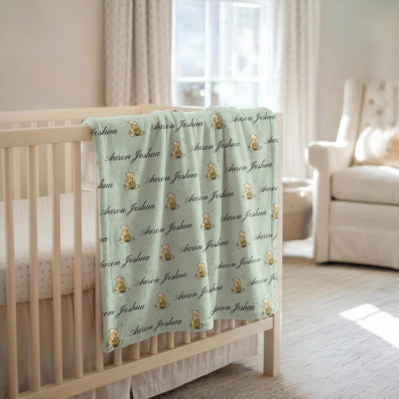 May include: A light green baby blanket draped over a crib. The blanket features a repeating pattern of a teddy bear holding a honey pot, along with the name "Aaron Joshua" in a script font. The blanket is made of soft material.
