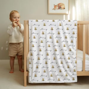 May include: White baby blanket with a repeating pattern of a teddy bear, stars, and the name "Avery" in a script font. The blanket is draped over a wooden crib. A framed picture of a fox is on the wall.