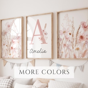 May include: Three framed floral art prints. The prints feature watercolor illustrations of pink flowers and foliage. The center print has a large pink letter "A" with the name "Amelia" below. The frames are light wood. The text "MORE COLORS" is at the bottom.