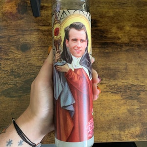May include: A glass prayer candle featuring a portrait of a man in religious attire. The candle has a gold and red design with a floral pattern at the base. The candle is held by a person.