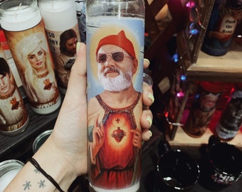 May include: A white candle with a red, white, and blue image of a man wearing a red hat and a beard. The man is holding a red heart with a cross on it. The candle is in a glass jar.