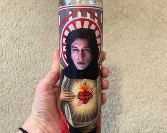 May include: A white candle with a red, white, and black image of a man with a heart and two crossed lightsabers. The image is surrounded by a red and white circle.