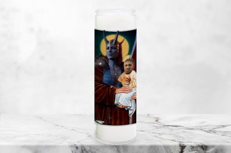 May include: A white candle with a blue and red image of a woman holding a child. The woman has blue skin and horns. The child has a halo and is wearing a white robe. The candle is decorated with gold accents.