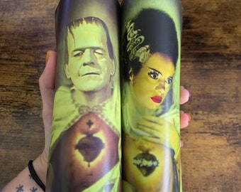 May include: Two yellow candles with images of Frankenstein's monster and his bride. The candles are in glass jars with a green background.