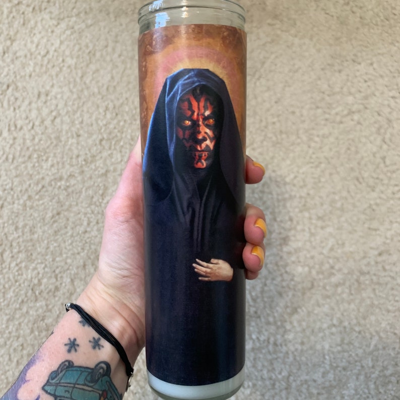 May include: A red and black Darth Maul candle with a gold halo. The candle is in a glass jar with a white label.