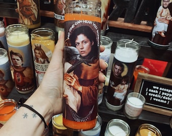 May include: A glass candle with an orange wax base and a printed image of a woman holding a baby. The image is from the movie Alien. The text on the candle reads "No one can hear you scream".