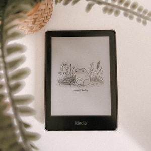 May include: A black Kindle e-reader displaying a whimsical illustration of a frog reading in a field, with the text "toadally booked." The Kindle is on a white surface, with a woven basket and greenery in the background.