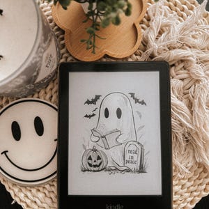 May include: A black and white illustration of a ghost reading a book on a Kindle e-reader. The ghost is sitting next to a jack-o-lantern and a tombstone that reads "read in peace".