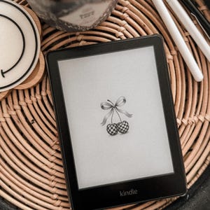 Puede incluir: Un lector electrónico Kindle en blanco y negro con un diseño de cereza a cuadros en la pantalla. El lector electrónico está sobre una bandeja de mimbre tejido.