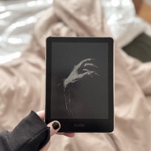 May include: A black and white illustration of a hand reaching out from the darkness on a Kindle e-reader. The e-reader is black with the word "kindle" in white text on the bottom.