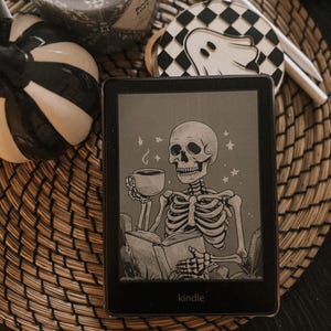 May include: A black and white illustration of a skeleton holding a cup of coffee and reading a book on a Kindle e-reader. The background is a gray gradient with stars.