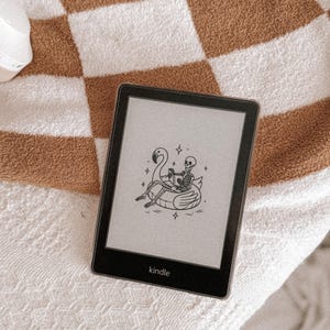 Puede incluir: Lector de libros electrónicos Kindle negro con una ilustración de un esqueleto en un flotador de flamenco. El lector está sobre una toalla a cuadros blancos y marrones. El logotipo de Kindle es visible.