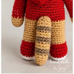 May include: A handmade crochet doll with a red body, tan legs, and a striped tail. The doll's feet are red with white soles. The text "handmade by Joy" is written in cursive on the bottom right corner.