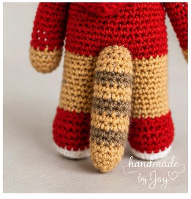 May include: A handmade crochet doll with a red body, tan legs, and a striped tail. The doll's feet are red with white soles. The text "handmade by Joy" is written in cursive on the bottom right corner.
