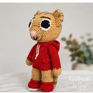 May include: A handmade crochet tiger doll wearing a red hoodie and shorts. The doll has large black and white eyes, a pink nose, and tan and brown striped details. The doll is standing on a white surface. The text "handmade by Joy" is in the bottom right corner.