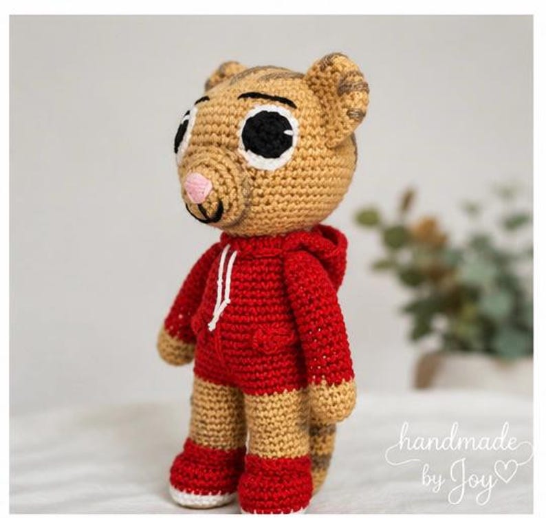 May include: A handmade crochet tiger doll wearing a red hoodie and shorts. The doll has large black and white eyes, a pink nose, and tan and brown striped details. The doll is standing on a white surface. The text "handmade by Joy" is in the bottom right corner.