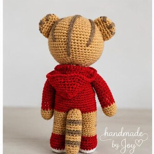 May include: A handmade crochet tiger toy, viewed from the back, wearing a red hooded sweatshirt. The tiger has tan and brown stripes, with a matching tail. The text "handmade by Joy" is written in cursive.