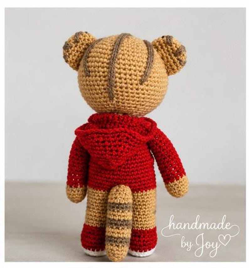 May include: A handmade crochet tiger toy, viewed from the back, wearing a red hooded sweatshirt. The tiger has tan and brown stripes, with a matching tail. The text "handmade by Joy" is written in cursive.