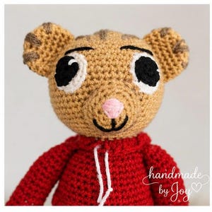 May include: A handmade crochet tiger doll wearing a red hoodie. The tiger has tan fur with brown stripes on its ears, black eyes, a pink nose, and a smiling mouth. The text "handmade by Joy" is written in white script.