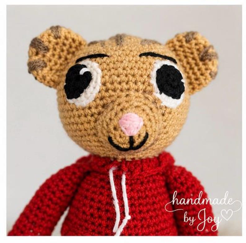 May include: A handmade crochet tiger doll wearing a red hoodie. The tiger has tan fur with brown stripes on its ears, black eyes, a pink nose, and a smiling mouth. The text "handmade by Joy" is written in white script.