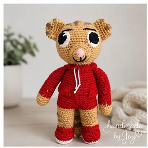 May include: A handmade crochet tiger doll wearing a red hoodie and shorts. The tiger has tan and brown fur with black eyes, a pink nose, and a smiling mouth. The doll is approximately 25 cm tall.