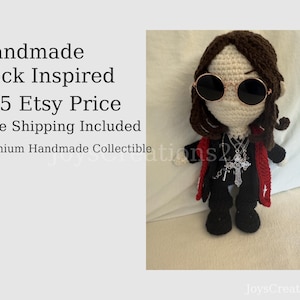 May include: Handmade crochet doll inspired by rock music. The doll has brown hair, round sunglasses, and a black outfit with a red vest. It also has a silver cross necklace. The text "Handmade Rock Inspired" is visible.