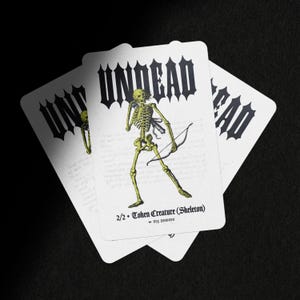 May include: A white rectangular game card featuring a yellow skeleton illustration holding a bow and arrow. The text UNDEAD is printed in a bold black gothic font at the top, with 2/2 Token Creature (Skeleton) and ms brown at the bottom.