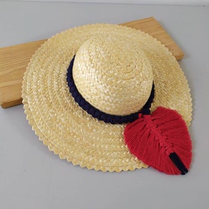 Peut inclure: Un chapeau de soleil en paille à large bord et un ruban bleu marine autour de la couronne. Un pompon rouge pend du ruban.