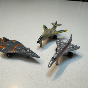 Op de afbeelding: Drie speelgoedvliegtuigen, een grijs en oranje gevechtsvliegtuig, een zilveren en rode Concorde en een bruine en blauwe bommenwerper.
