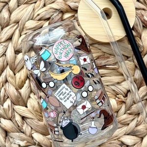 May include: A clear glass can-shaped tumbler with a bamboo lid and two straws. The tumbler is decorated with colorful stickers featuring a wizarding theme, including text that reads "Happee Birthdae Harry". The tumbler is sitting on a woven surface.