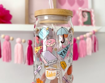 May include: A clear glass tumbler with a bamboo lid and a glass straw. The tumbler is decorated with colorful stickers including a donut, umbrella, and a coffee cup that says "Luke's". The tumbler is held in a hand.