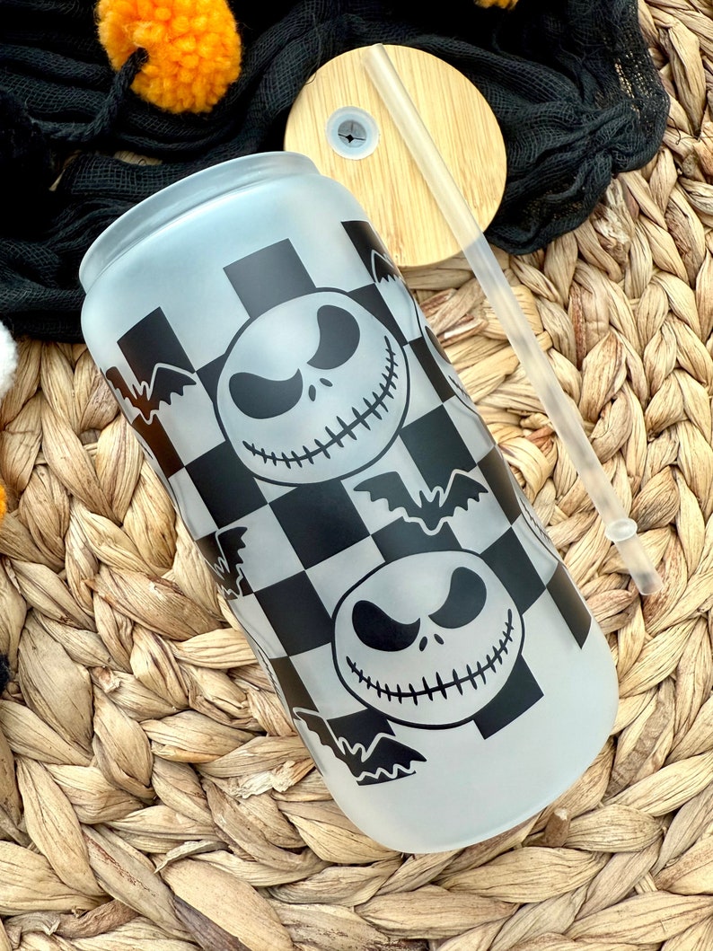May include: A frosted glass water bottle with a black and white checkered pattern and a repeated image of Jack Skellington's face from the Nightmare Before Christmas. The bottle has a black lid and a clear straw.
