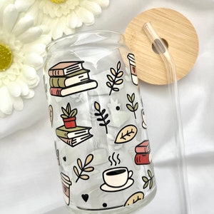 May include: A clear glass reusable can with a straw and lid. The can has a pattern of books, coffee cups, plants, and leaves. The lid is made of bamboo.